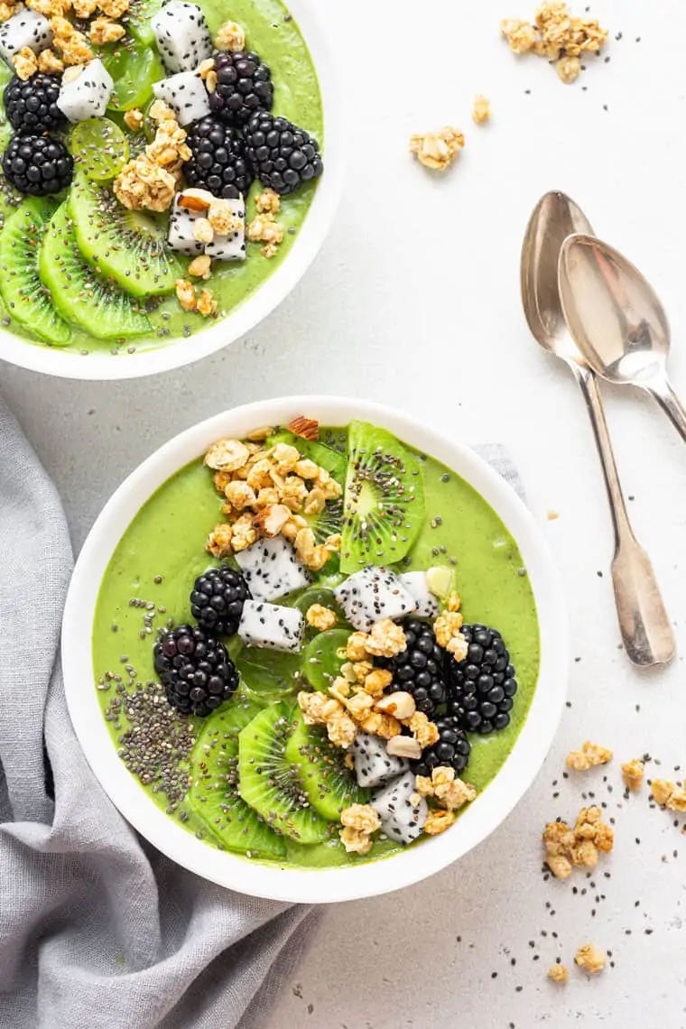 overhead of two white bowls of green smoothie bowl with fresh fruit and berries on top
