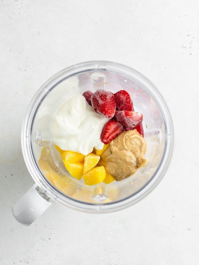 Make this strawberry mango smoothie with dairy-free yogurt, nut butter, chia seeds, and frozen fruit for a filling, high-protein breakfast. Vegan strawberry mango smoothie ingredients inside the blender.