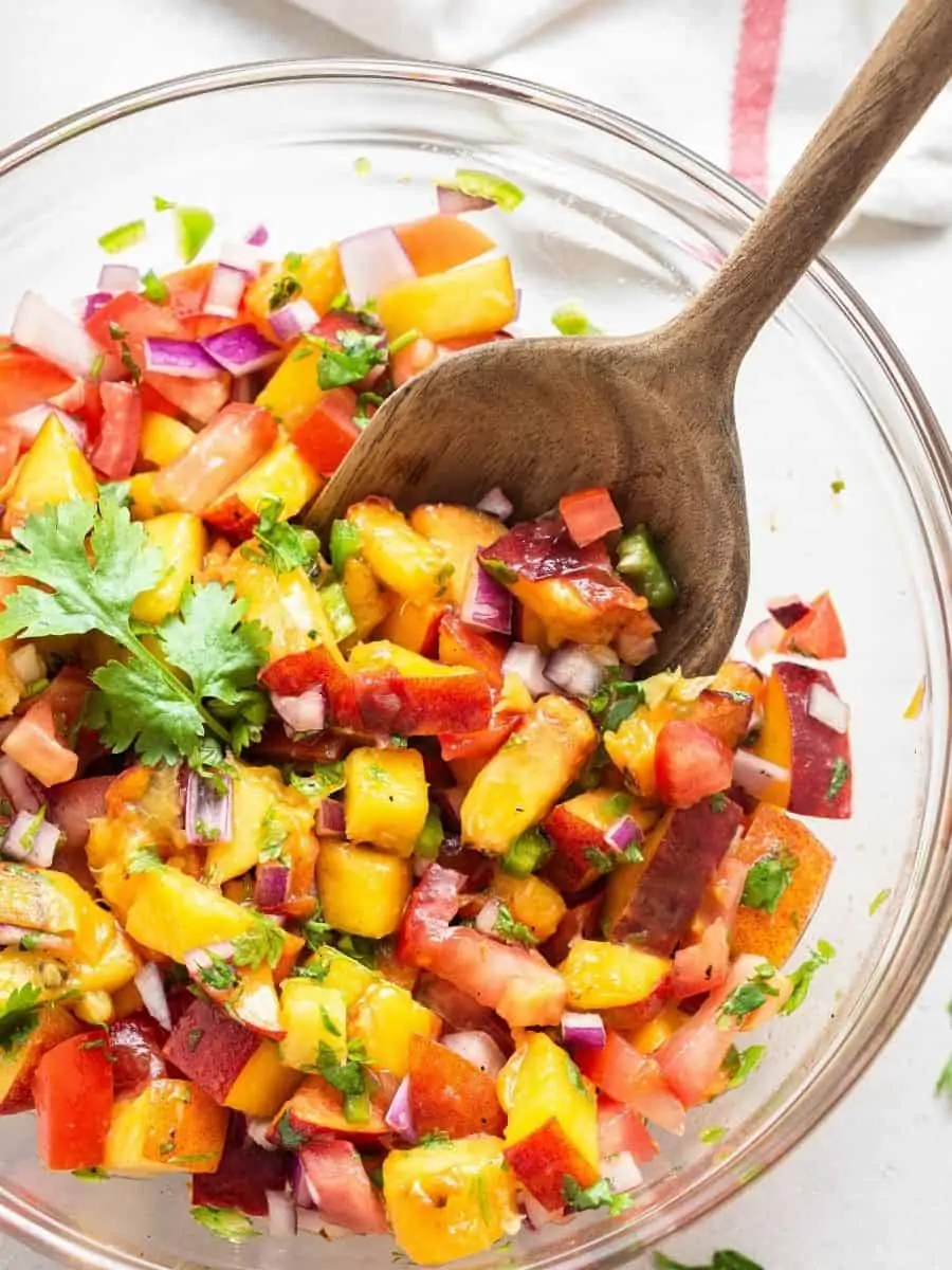This refreshing Peach Salsa recipe is a sweet, savory and spicy homemade condiment you’ll love with fish, chips and more. Easy and delicious! Overhead view of peach salsa in bowl with wooden spoon