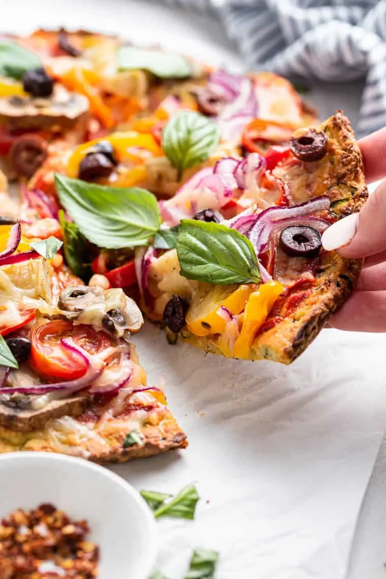 hand holding a piece of pizza with basil and olives