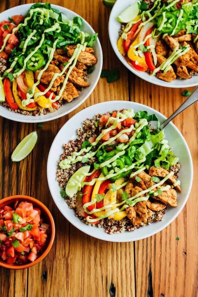 Overhead view of three white bowls with chicken, quinoa and fajita veggies served with a drizzle of green sauce.