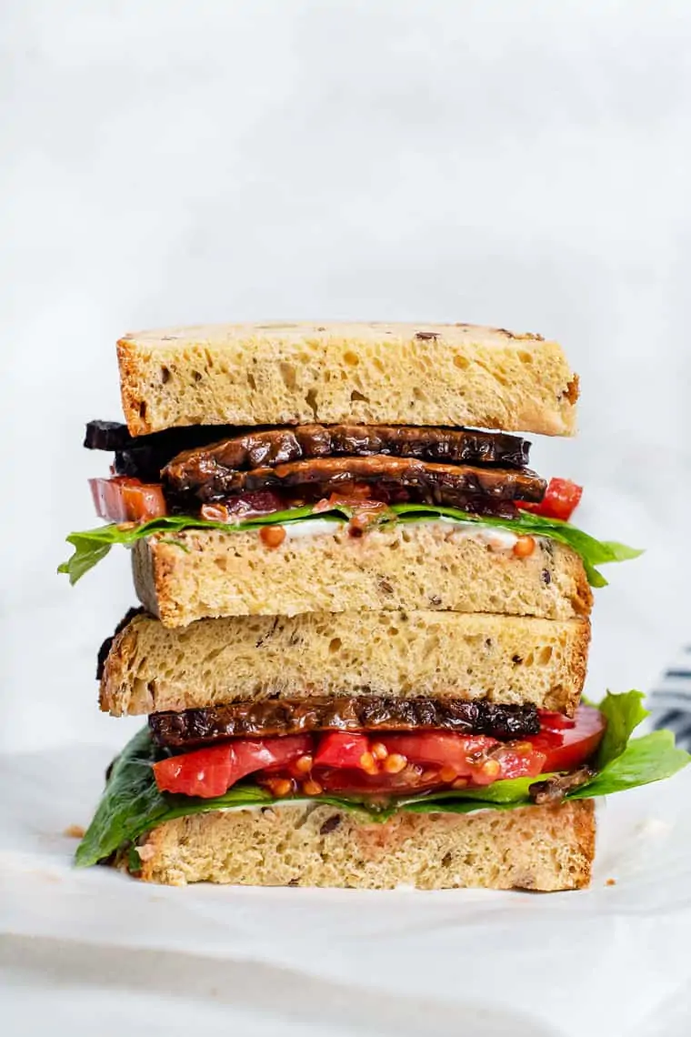 Learn how to make crispy, smoky tempeh bacon at home! This easy vegan recipe is perfect for salads, BLT sandwiches, breakfast, and more. Two sandwich halves with tempeh bacon stacked on top of one another.