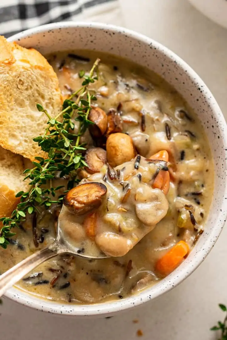 Close up of a spoonful of wild rice soup above a bowl of soup, garnished with bread and thyme sprigs. 