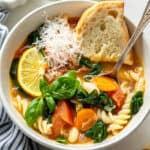 Overhead view of vegetable stew in bowl with Parmesan, lemon slice, basil, and bread