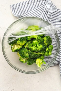 peeling plastic wrap off of a bowl of cooked broccoli