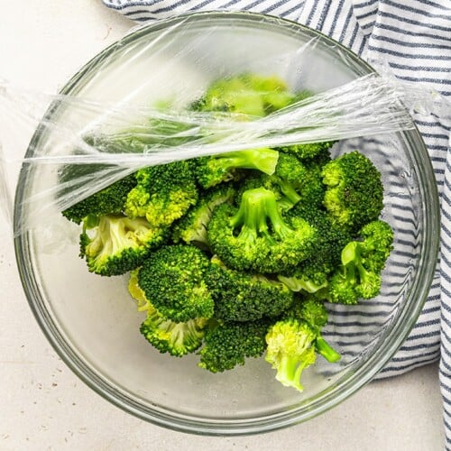 peeling plastic wrap off of a bowl of cooked broccoli