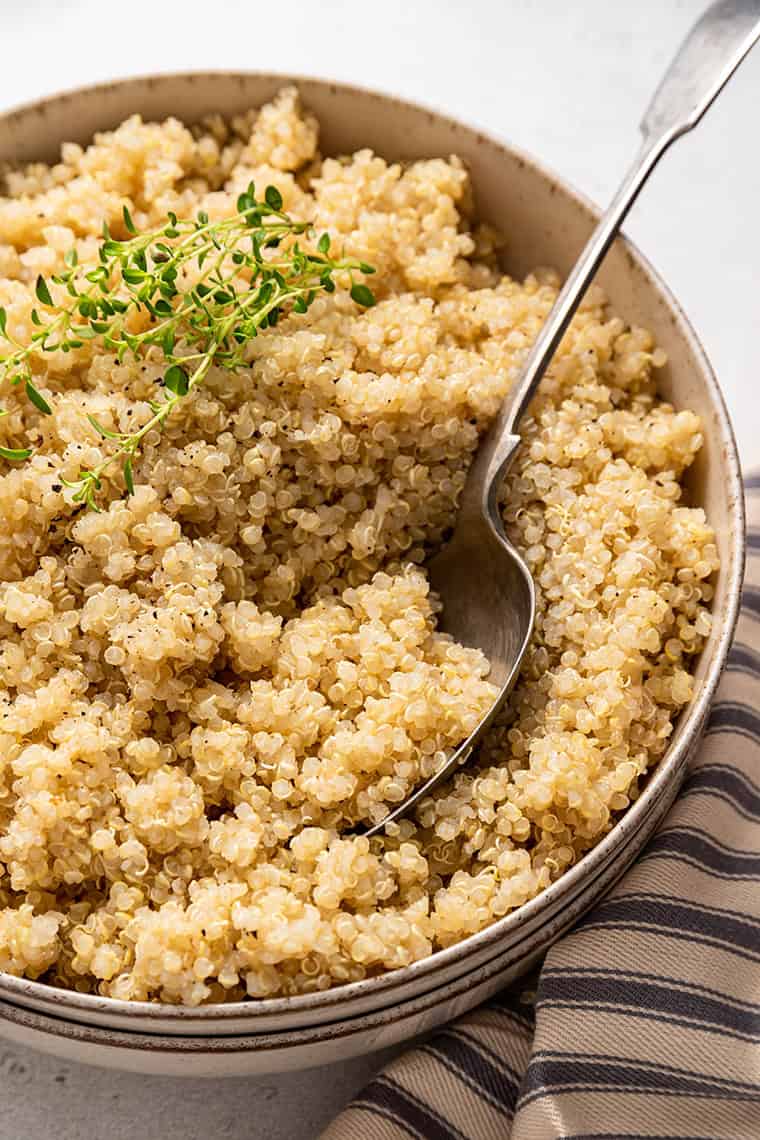 Everything you need to know about how to cook quinoa, including the amount of water, the different methods, and how healthy it is. Close up of a bowl of quinoa with a sprig of thyme on top, with a spoon in it