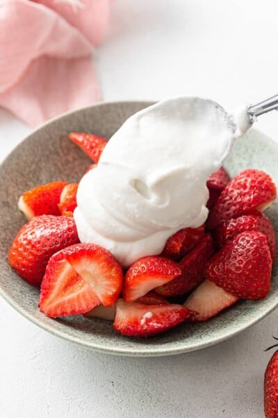 A spoon dolloping fluffy coconut whipped cream onto a bowl of strawberries.