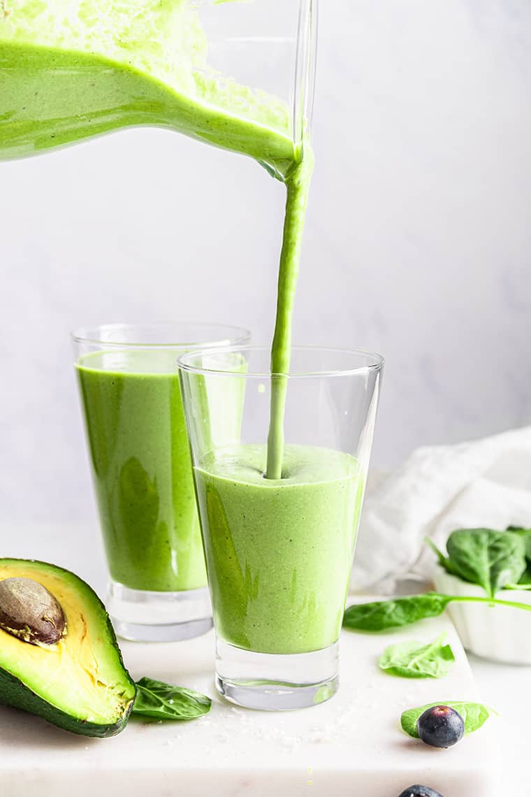 Pouring green smoothie from pitcher into glass, with ingredients scattered in foreground