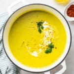 A pot of broccoli soup topped with vegan creme fraiche, broccoli, olive oil, and chili flakes, next to a ramekin of chili flakes and a jar of garlic oil