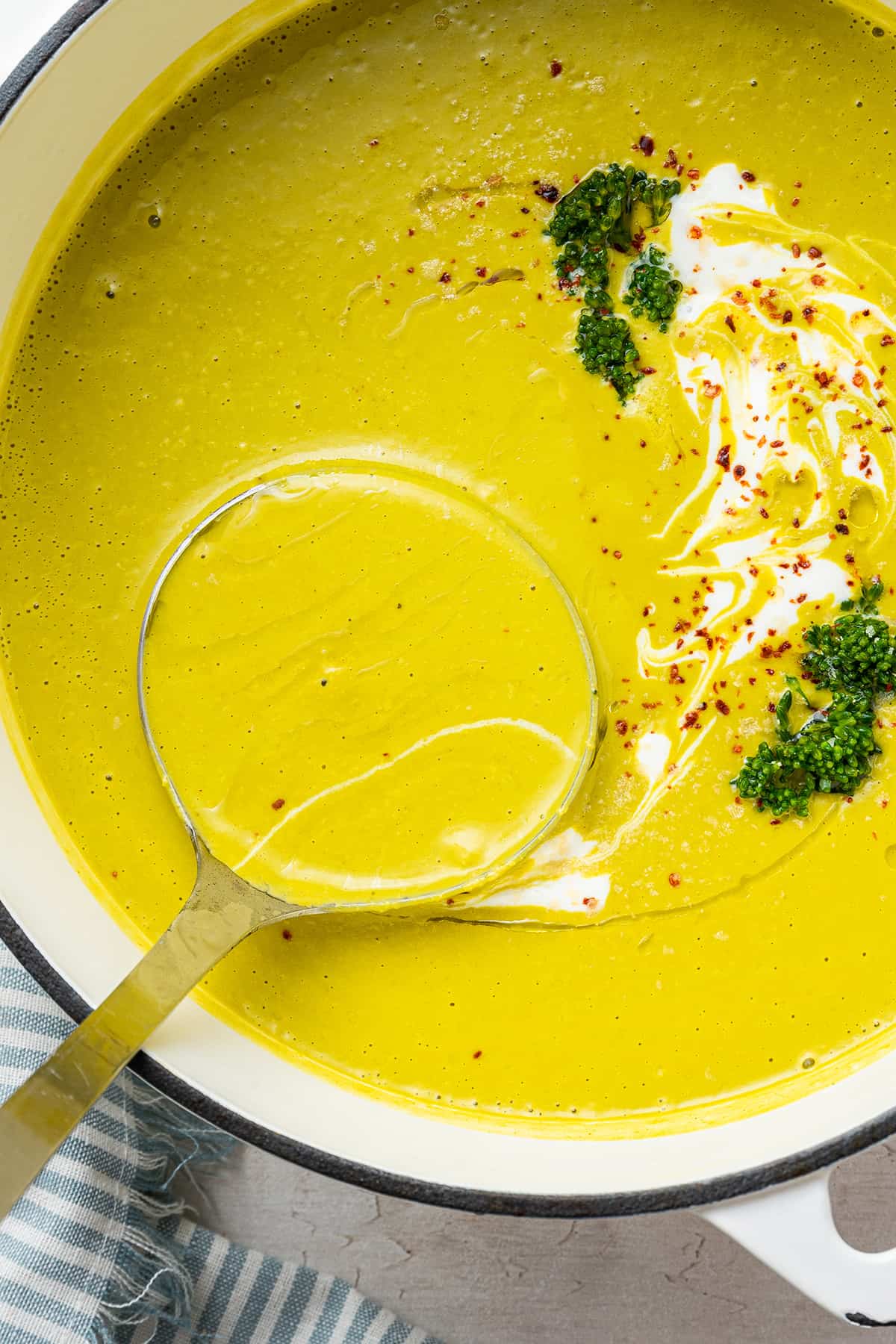 A ladle scooping out a spoonful of broccoli soup from a pot of broccoli soup garnished with chili flakes, broccoli, and vegan creme fraiche
