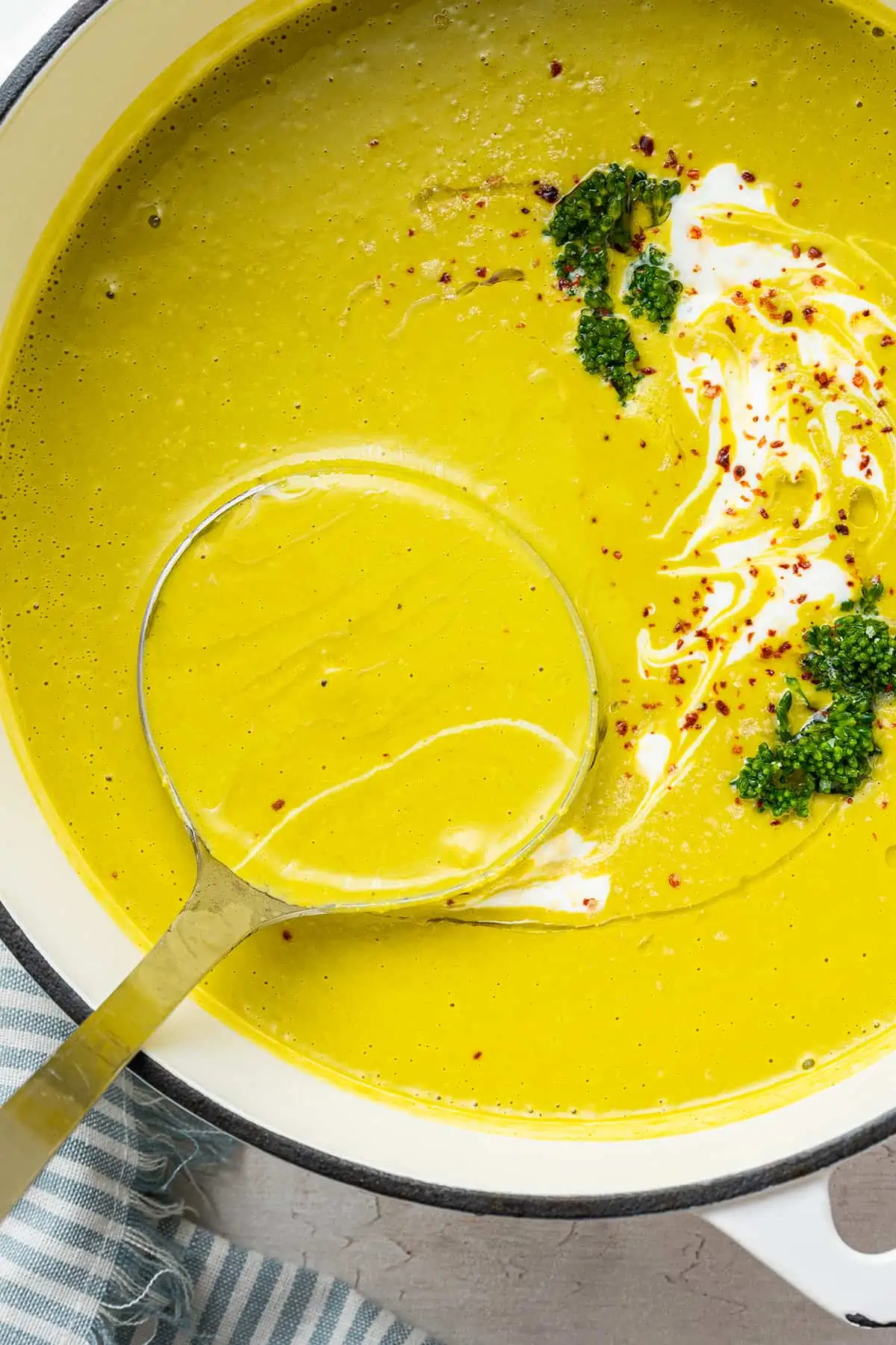 A ladle scooping out a spoonful of broccoli soup from a pot of broccoli soup garnished with chili flakes, broccoli, and vegan creme fraiche