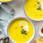 Overhead view of two bowls of broccoli soup garnished with a lemon slice, broccoli, vegan creme fraiche, and chili flakes, next to a pot of soup and some spoons