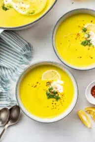 Overhead view of two bowls of broccoli soup garnished with a lemon slice, broccoli, vegan creme fraiche, and chili flakes, next to a pot of soup and some spoons