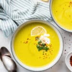 Overhead view of a bowl of broccoli soup garnished with broccoli, chili flakes, vegan creme fraiche, and a lemon slice, next to another bowl, some lemon slices, and some spoons
