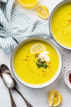 Overhead view of a bowl of broccoli soup garnished with broccoli, chili flakes, vegan creme fraiche, and a lemon slice, next to another bowl, some lemon slices, and some spoons