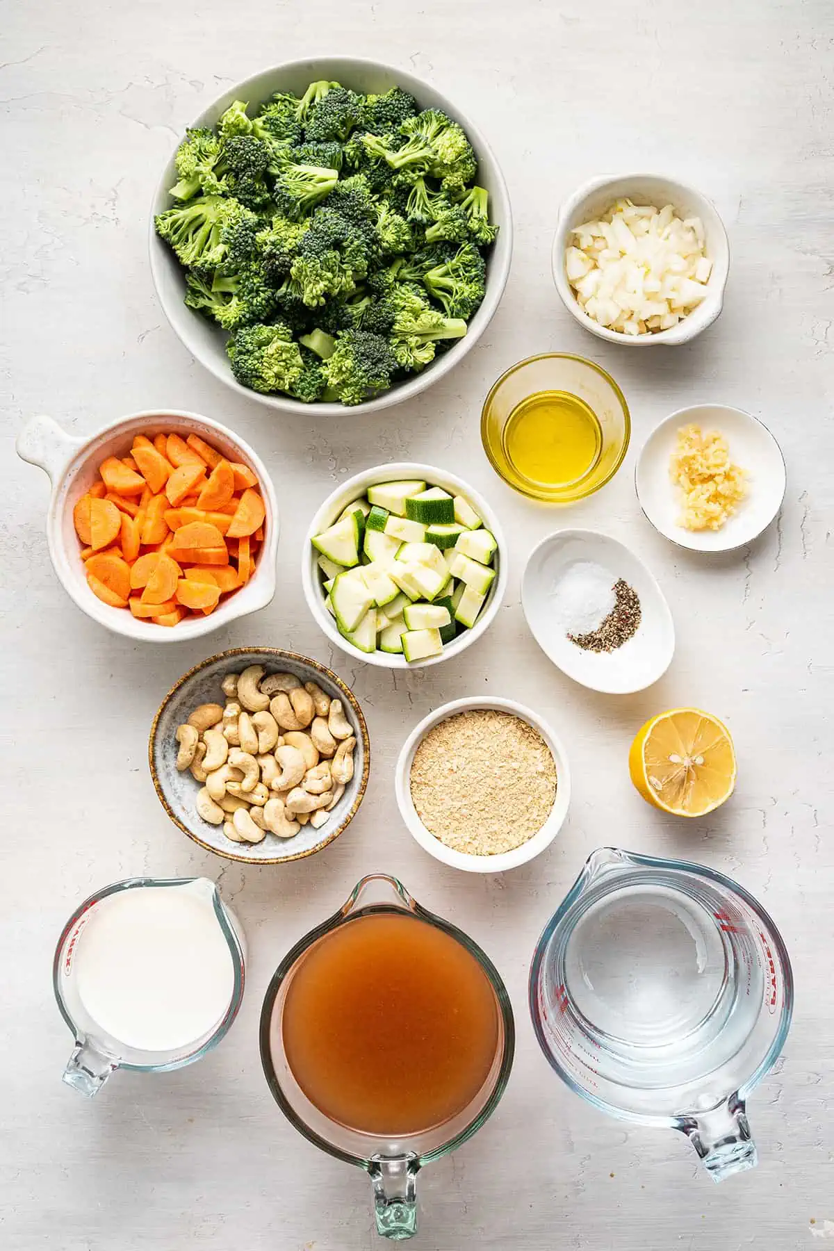 Overhead view of the ingredients needed for vegan cream of broccoli soup: A bowl of broccoli florets, a bowl of diced carrots, a bowl of diced onions, a bowl of diced zucchini, a bowl of raw cashews, a glass of almond milk, a glass of vegetable broth, a glass of water, a ramekin of salt and pepper, a ramekin of olive oil, a ramekin of chopped garlic, a ramekin of nutritional yeast, and half a lemon