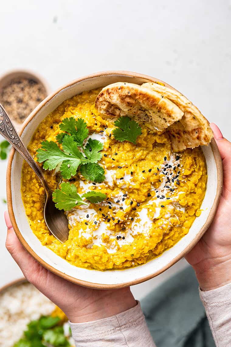 Two hands holding bowl of red lentil dal with spoon, flatbread, and cilantro garnish