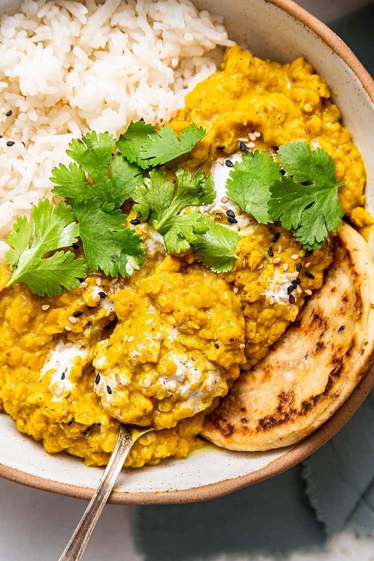 Overhead view of red lentil dal in bowl with spoon and white rice, flatbread, and cilantro