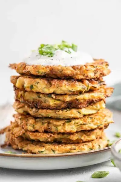 Stack of crispy zucchini fritters on plate, topped with yogurt and scallions