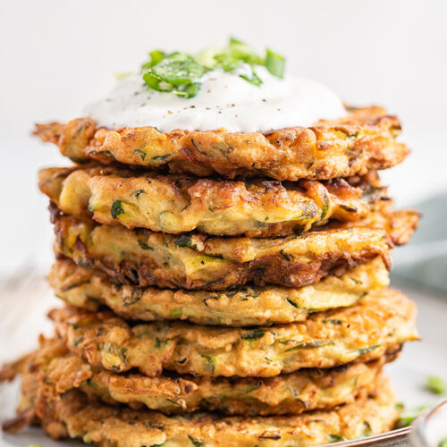 Stack of crispy zucchini fritters on plate, topped with yogurt and scallions