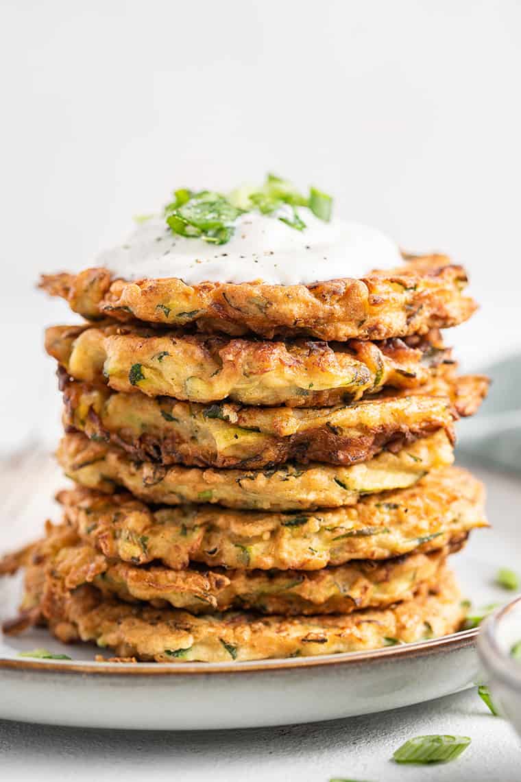 Stack of crispy zucchini fritters on plate, topped with yogurt and scallions