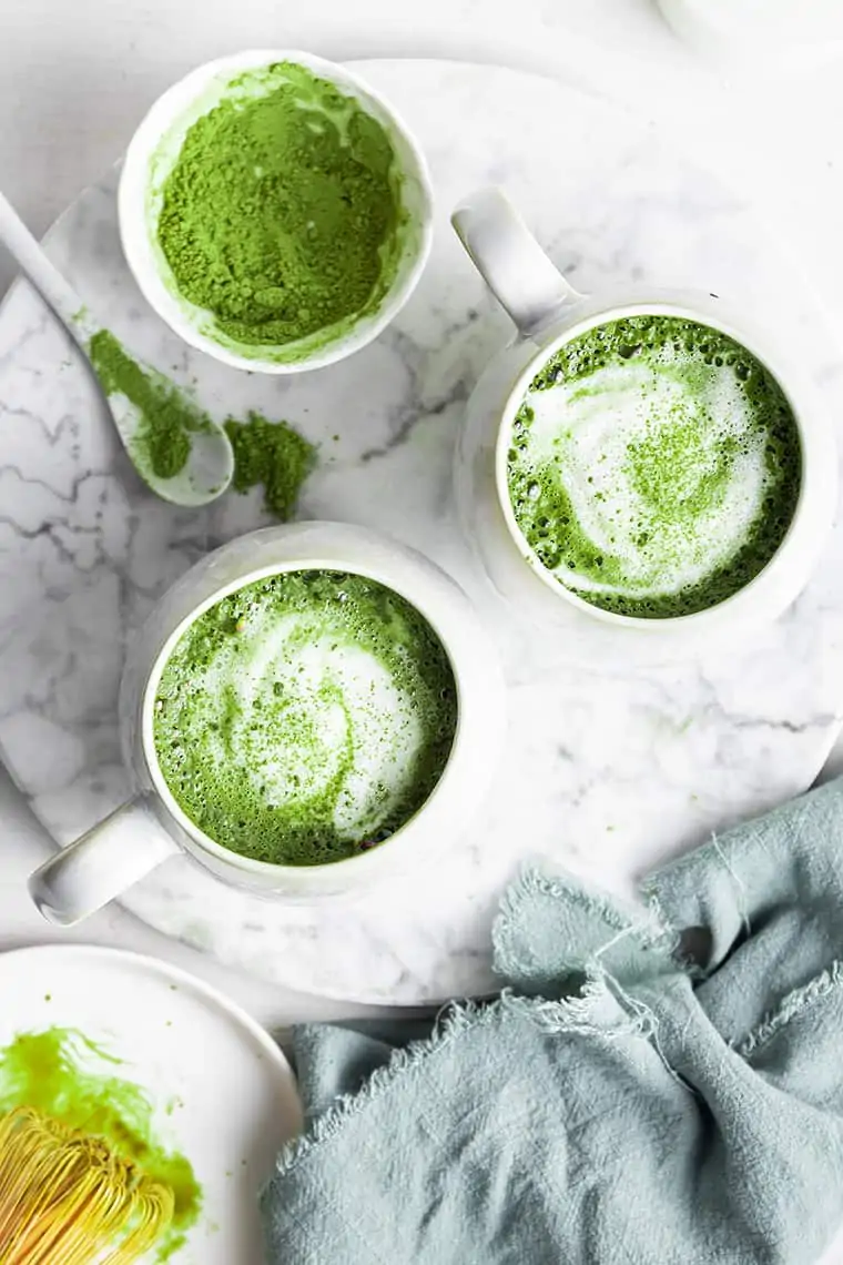 Overhead view of two mugs of matcha latte with matcha powder in small bowl