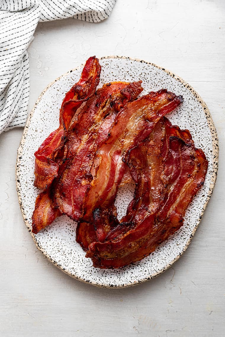 How to make air fryer bacon, which is crispy, juicy, and more convenient than any other method of cooking bacon. A white oval plate with a pile of cooked bacon on it
