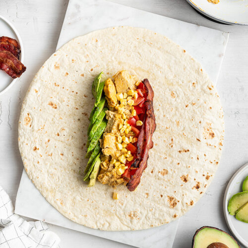 A tortilla with a row of bacon, a row of avocado, and a row of eggs on it, topped with veggies
