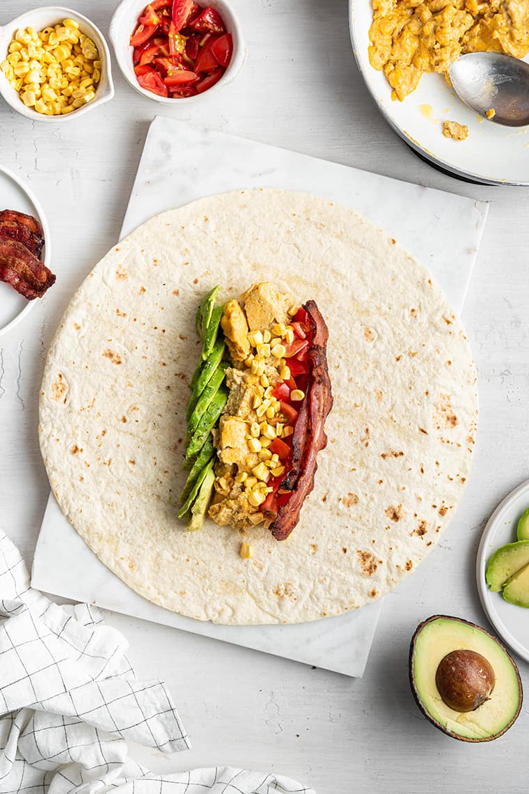 A tortilla with a row of bacon, a row of avocado, and a row of eggs on it, topped with veggies