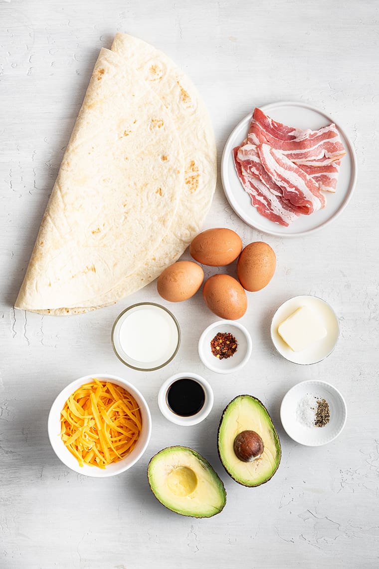 A folded tortilla next to a plate of uncooked bacon, four whole eggs, an avocado cut in half, a bowl of shredded cheese, a bowl of chili flakes, a knob of butter, a bowl of milk, a bowl of soy sauce, and a bowl of salt and pepper