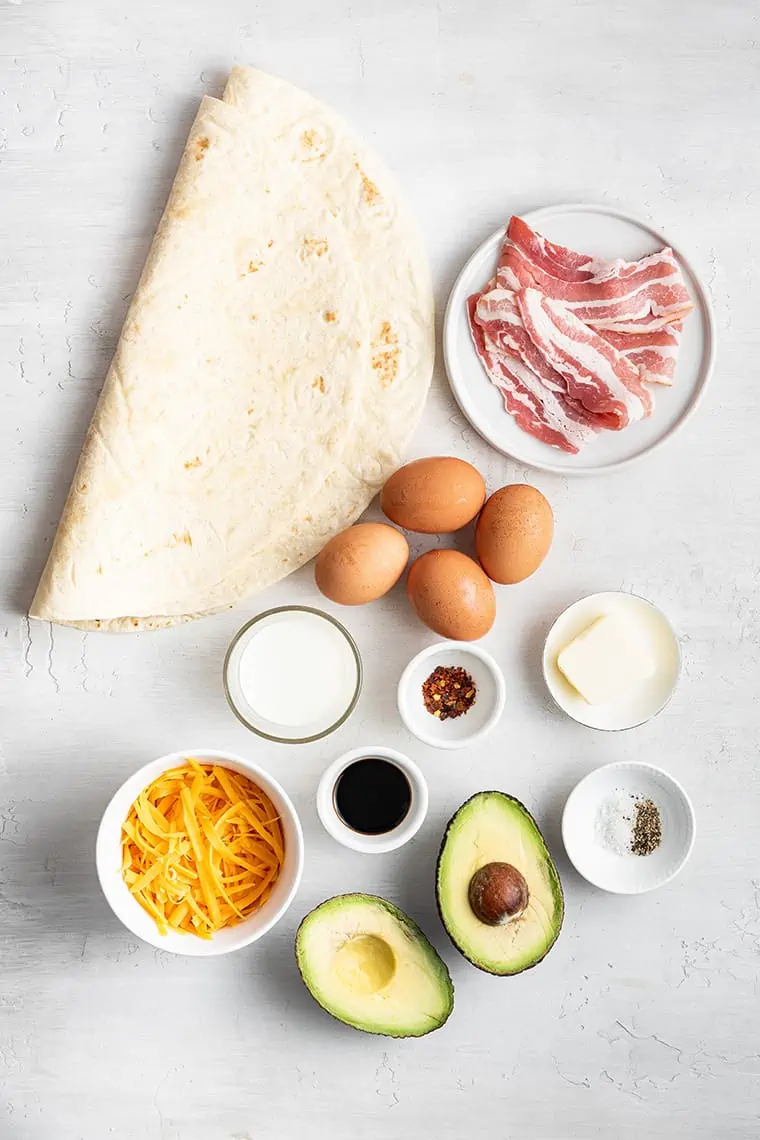 A folded tortilla next to a plate of uncooked bacon, four whole eggs, an avocado cut in half, a bowl of shredded cheese, a bowl of chili flakes, a knob of butter, a bowl of milk, a bowl of soy sauce, and a bowl of salt and pepper