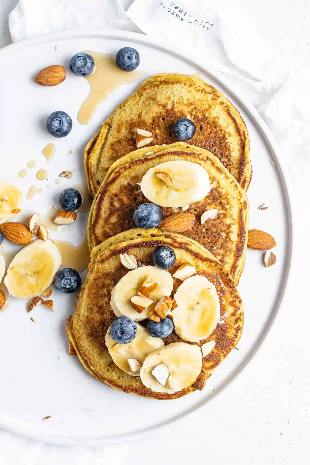 These protein pancakes are fluffy and flavorful, made with 6 ingredients in the blender. Gluten-free, dairy-free, and meal prep-friendly! Top view of three protein pancakes on a white plate topped with fruit and chopped nuts.