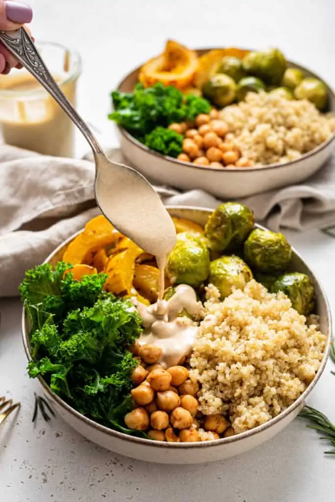 A spoon drizzles tahini dressing over an autumn harvest bowl.