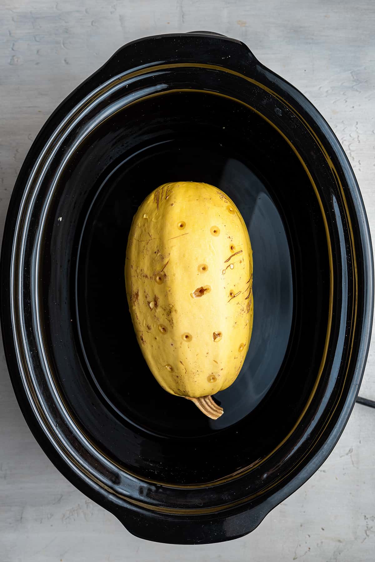 A whole spaghetti squash, with holes poked in it, inside a slow cooker