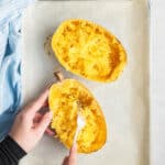 A cooked spaghetti squash, cut in half, on a baking tray covered in parchment paper, with a hand using a fork to shred it