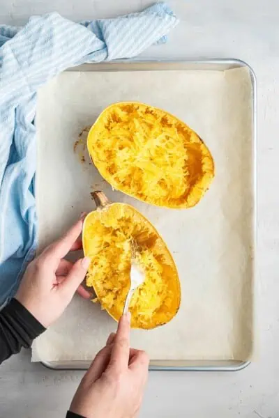 A cooked spaghetti squash, cut in half, on a baking tray covered in parchment paper, with a hand using a fork to shred it
