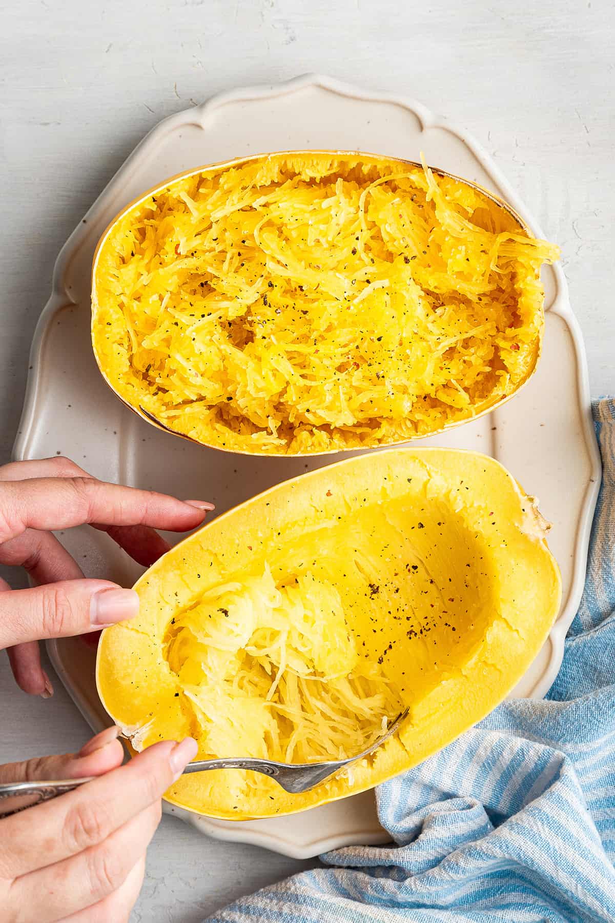 A cooked spaghetti squash cut in half on a serving tray, with one half shredded, and the other half in the process of being shredded by a fork