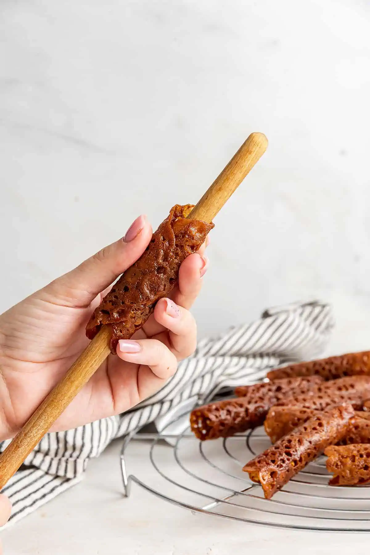 A hand pulling a shaped brandy snap cookie off of a wooden spoon handle, with lots of cookies in the background