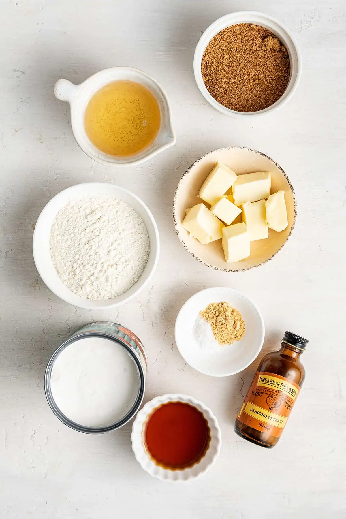 Overhead view of the ingredients for brandy snaps: a bowl of vegan butter, a bowl of sugar, a bowl of agave, a bowl of flour, a bowl of coconut cream, a bowl of seasonings, a bowl of maple syrup, and a bottle of vanilla extract