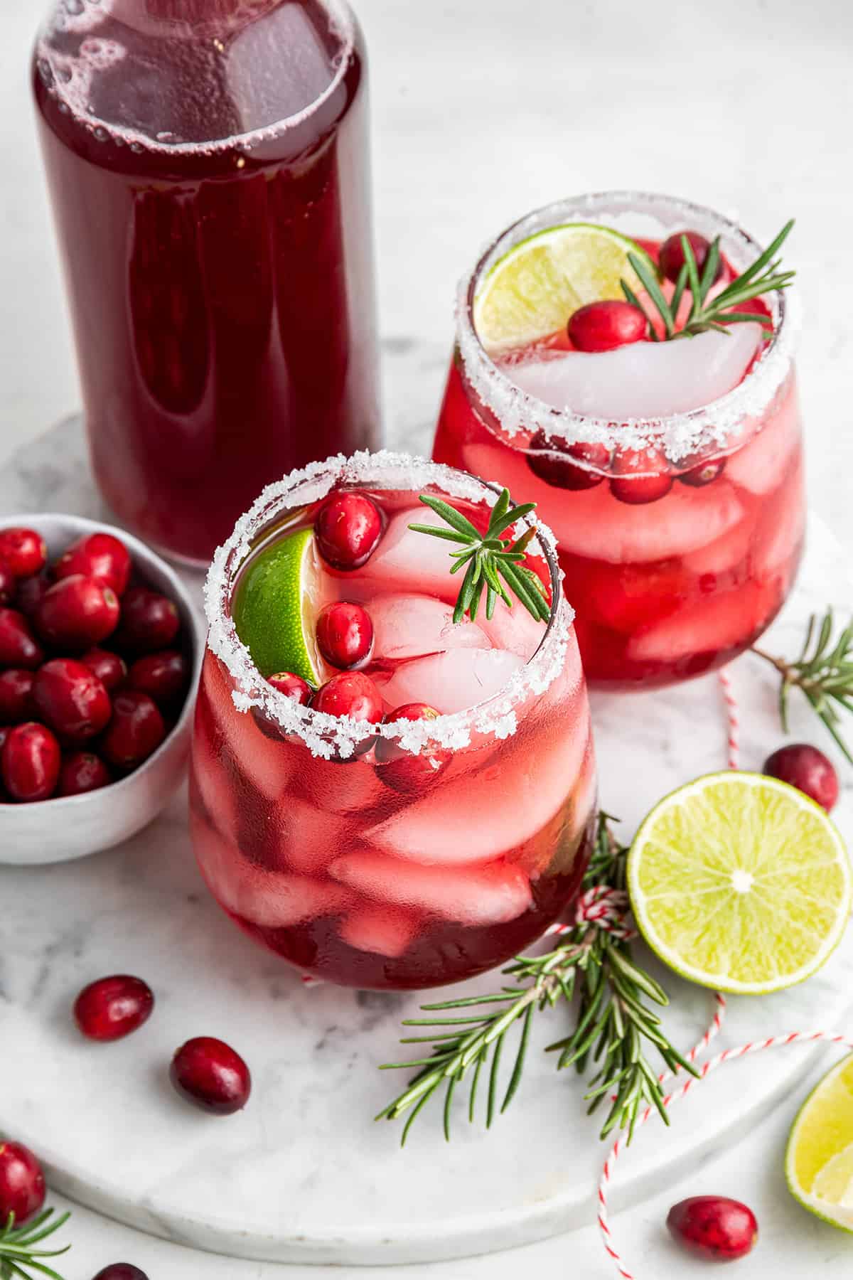 Two cranberry margaritas topped with fresh cranberries, lime wedges, and fresh rosemary, next to a bowl of cranberries, a bottle of cranberry juice, some sprigs of rosemary, and some lime halves