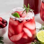 A cranberry margarita in a salt-rimmed glass, surrounded by rosemary sprigs, cranberries, and lime wedges