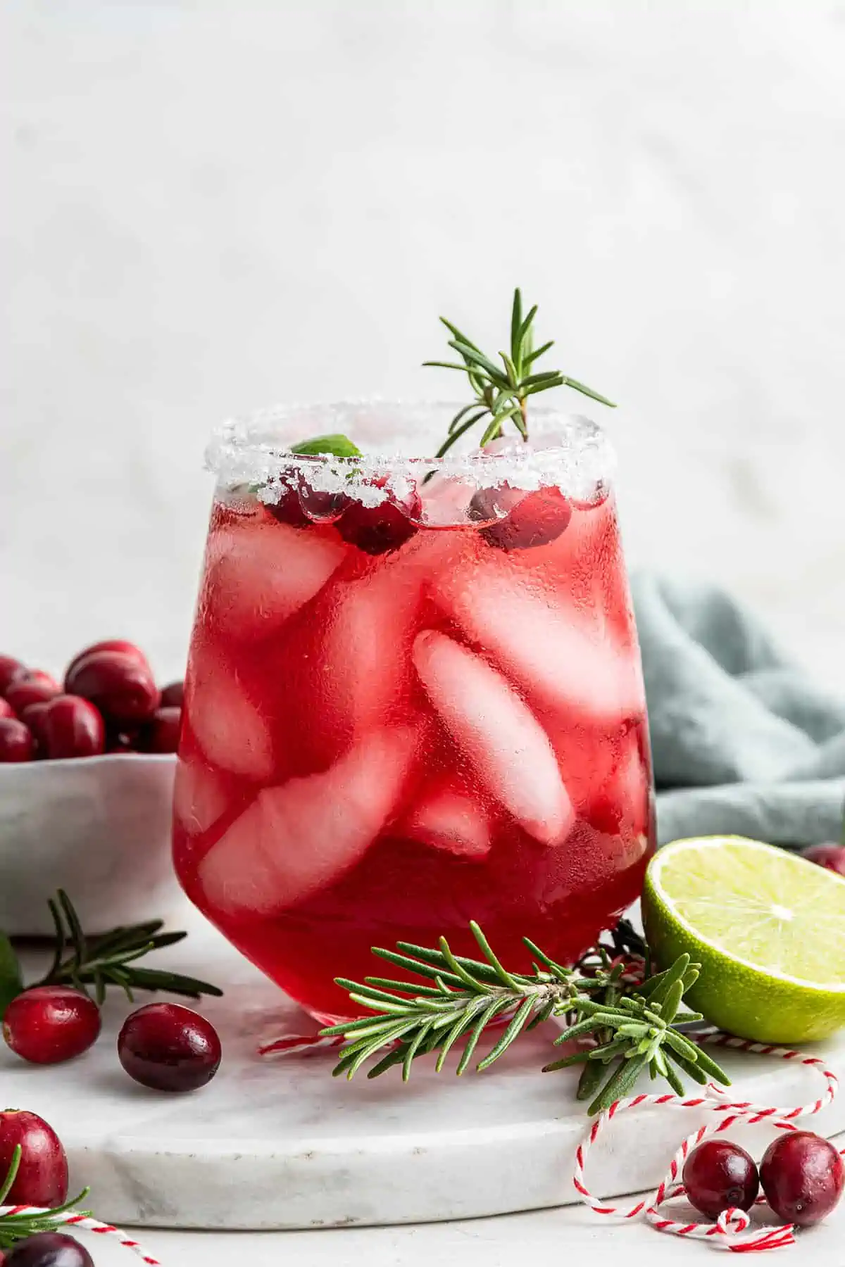 A cranberry margarita with a sprig of rosemary sticking out, next to a bowl of cranberries, half a lime, and two sprigs of rosemary