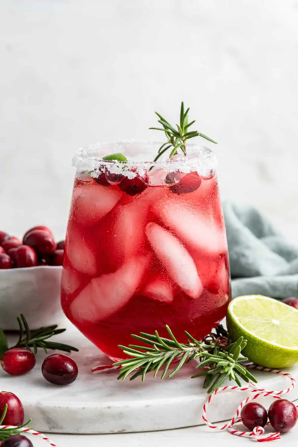 This 5-minute cranberry margaritas recipe is tart and sweet, has a nice kick from tequila, and is the perfect drink with chips and salsa. A cranberry margarita with a sprig of rosemary sticking out, next to a bowl of cranberries, half a lime, and two sprigs of rosemary