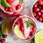 Overhead view of two cranberry margaritas, topped with lime wedges, fresh cranberries, and rosemary sprigs, next to a bowl of cranberries and lime wedges.
