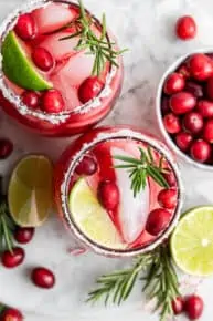 Overhead view of two cranberry margaritas, topped with lime wedges, fresh cranberries, and rosemary sprigs, next to a bowl of cranberries and lime wedges.