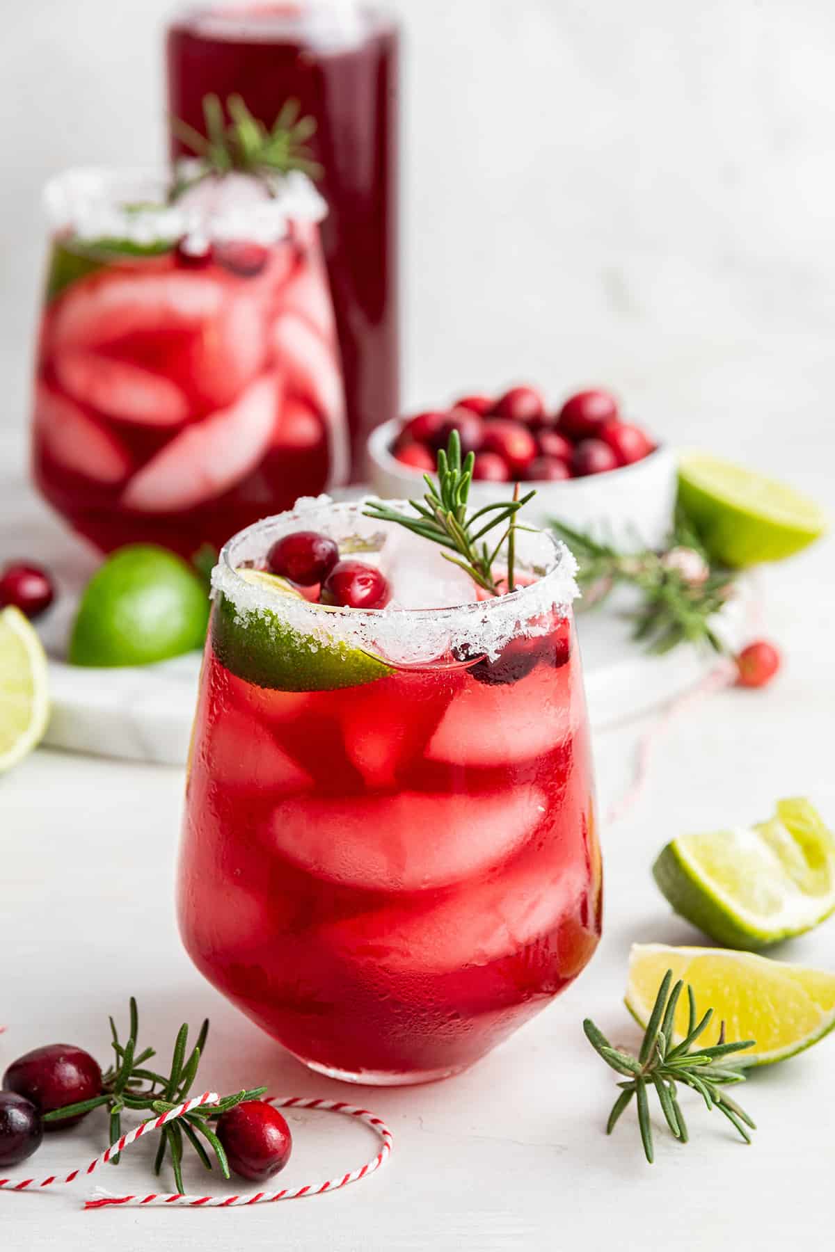 A cranberry margarita in a salt-rimmed glass, topped with a sprig of rosemary, with another cranberry margarita and a bowl of cranberries in the background, and lime wedges all around.