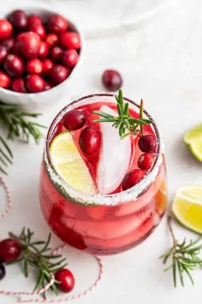 Overhead view of a cranberry margarita in a salt-rimmed glass, topped with a lime wedge, cranberries, and a sprig of rosemary, next to a bowl of cranberries and some rosemary sprigs