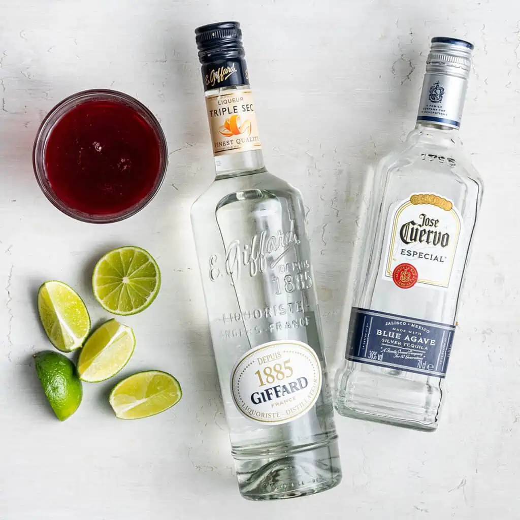 Overhead view of a bottle of tequila, a bottle of triple sec, a glass of cranberry juice, and a lime cut into wedges.