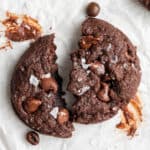 An espresso cookie broken in half, with chocolate smeared on the table, surrounded by espresso beans